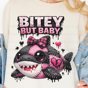 May include: Cream-colored sweatshirt featuring a graphic of a stitched shark with a pink bow and heart accents. The text "BITEY BUT BABY" is printed above the shark in a dripping font. The design incorporates black, pink, and white elements.