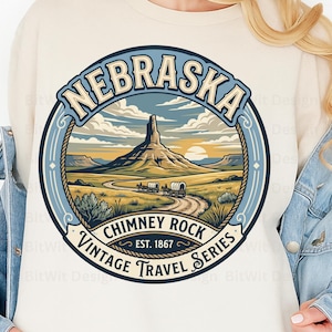 May include: A cream-colored sweatshirt featuring a circular graphic of Nebraska's Chimney Rock. The illustration depicts a landscape with a rock formation, a winding road, and covered wagons. Text reads: NEBRASKA, CHIMNEY ROCK, EST. 1867, VINTAGE TRAVEL SERIES.