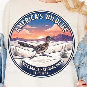 Peut inclure: Un sweat-shirt crème avec un motif circulaire représentant un grand géocoucou dans un paysage désertique. Le texte indique "AMERICA'S WILDLIFE", "WHITE SANDS NATIONAL PARK" et "EST. 1933". L'image montre des dunes blanches, des montagnes et un coucher de soleil.