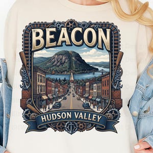 May include: A cream-coloured sweatshirt featuring a graphic print of a town street leading towards a mountain and river. The design includes the text BEACON and HUDSON VALLEY, framed by gears, anchors, and oars in shades of blue, brown, and tan.