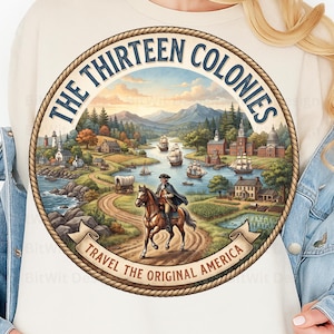May include: A cream-coloured sweatshirt featuring a circular graphic with a rope border. The text reads THE THIRTEEN COLONIES and TRAVEL THE ORIGINAL AMERICA. The illustration depicts a rider on horseback, sailing ships, colonial buildings, and mountains.