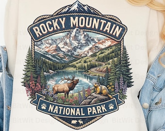 May include: A cream-colored sweatshirt featuring a graphic of Rocky Mountain National Park. The design includes snow-capped mountains, a winding road, a lake, an elk, and a marmot, with the text Rocky Mountain National Park and paw prints.