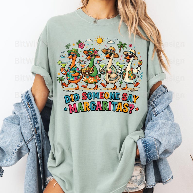 May include: A sage green t-shirt featuring a graphic of four ducks wearing hats and sunglasses, holding margaritas and snacks. The text reads, DID SOMEONE SAY MARGARITAS? The design includes palm trees, a sun, and flowers in a colorful, vibrant style.