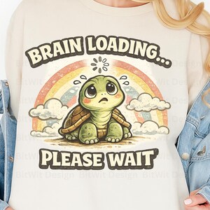 May include: A cream-coloured sweatshirt with a cartoon turtle illustration. The turtle has a worried expression, set against a rainbow and clouds. Text reads "BRAIN LOADING... PLEASE WAIT" above and below the turtle.