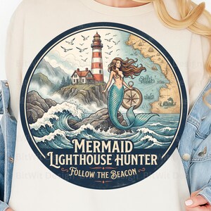 May include: A cream-coloured sweatshirt featuring a circular graphic of a mermaid holding a compass on a rocky shore near a lighthouse. The design includes a nautical map, crashing waves, and the text "MERMAID LIGHTHOUSE HUNTER FOLLOW THE BEACON".