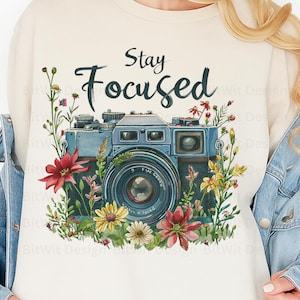 May include: Cream-colored sweatshirt featuring a blue vintage camera design surrounded by colorful flowers and foliage. The words "Stay Focused" are written above the camera in a script font. A stylish and artistic design.