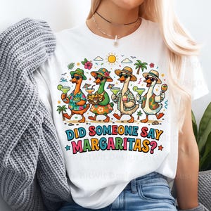 May include: A white t-shirt featuring a colorful graphic of four ducks wearing hats and sunglasses, holding margaritas and snacks under a sun and palm trees. The text below the ducks reads, DID SOMEONE SAY MARGARITAS? in bold, multi-colored letters.