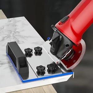 May include: A red power tool with a circular saw blade is positioned to cut a white marble surface. A black guide with blue trim and several black knobs are also visible, designed for precise cuts. The tool is designed for cutting stone or tile.
