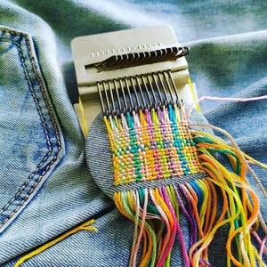 May include: A small, colorful woven textile project in progress, featuring a variety of bright threads in shades of yellow, green, pink, and blue. The weaving is attached to a denim patch, with a weaving tool in the background.
