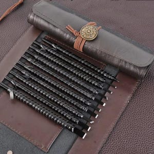 May include: A set of black metal sculpting tools with spiral handles, displayed in a brown leather roll-up case. The case is secured with a leather strap and a decorative bronze button. The tools are arranged neatly, showcasing their sharp edges.