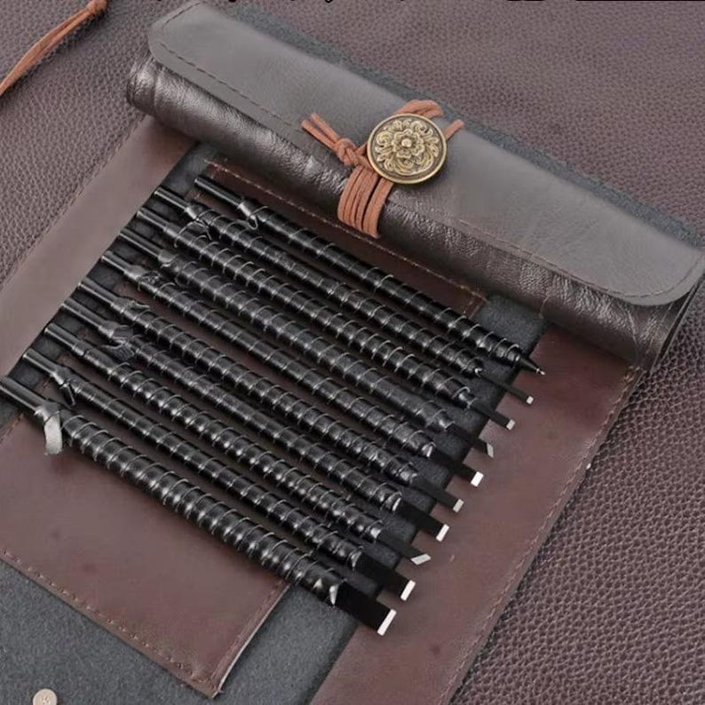 May include: A set of black metal sculpting tools with spiral handles, displayed in a brown leather roll-up case. The case is secured with a leather strap and a decorative bronze button. The tools are arranged neatly, showcasing their sharp edges.