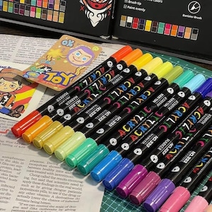 Puede incluir: Una colección de marcadores acrílicos en varios colores, dispuestos en diagonal. Cada marcador negro tiene la palabra "ACRYLIC" en letras coloridas. La imagen también incluye una pequeña tarjeta con un personaje de dibujos animados y la palabra "TOY".