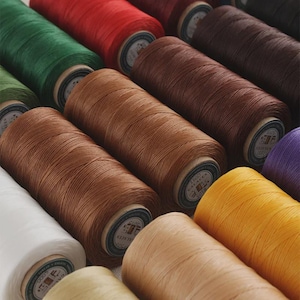 May include: A collection of cylindrical spools of thread in various colors including white, yellow, tan, brown, red, green, and purple. Each spool features a circular label on the end with the text 1500 GALACES and a logo.