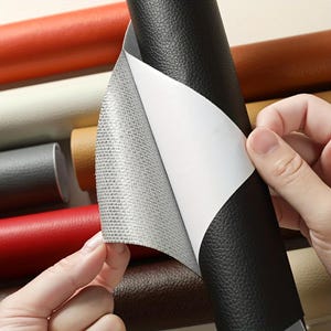 May include: A black self-adhesive faux leather sheet with a textured surface being peeled back to reveal a white paper backing and a grey fabric underside, with various other coloured rolls of faux leather visible in the background.