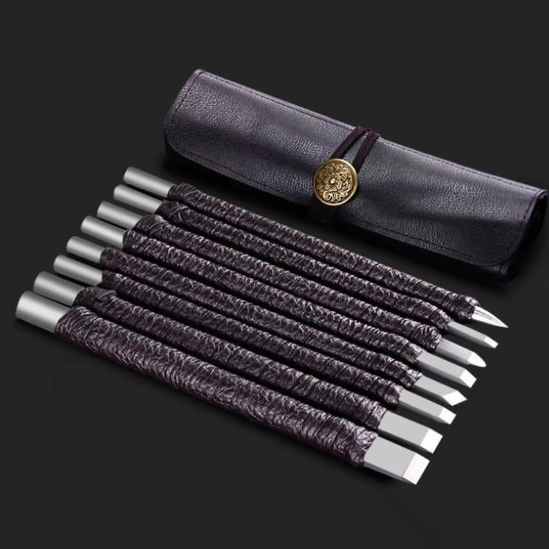 May include: A set of eight wood carving chisels with silver-colored metal tips and dark purple textured handles. The chisels are displayed with a dark purple leather tool roll with a decorative gold-colored button closure.