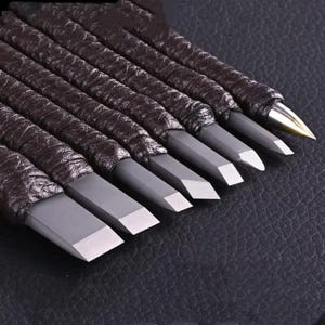 Pode incluir: Um conjunto de formões para marcenaria com cabos envoltos em couro marrom escuro. As lâminas dos formões são cinza com bordas de corte prateadas. As ferramentas estão dispostas diagonalmente, mostrando seu design afiado e preciso.
