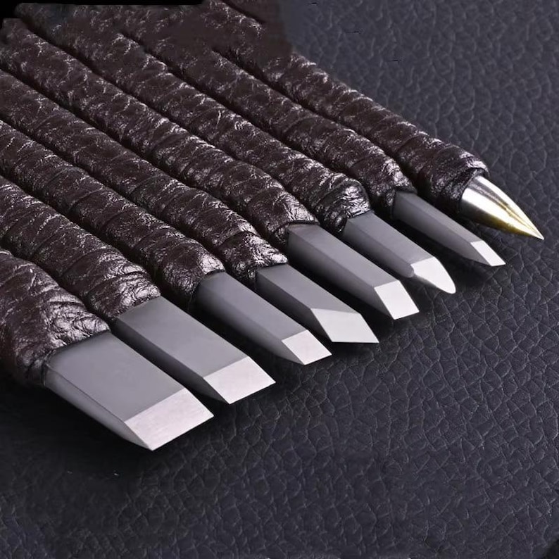 May include: A set of woodworking chisels with dark brown leather-wrapped handles. The chisel blades are gray with silver cutting edges. The tools are arranged diagonally, showcasing their sharp, precise design.