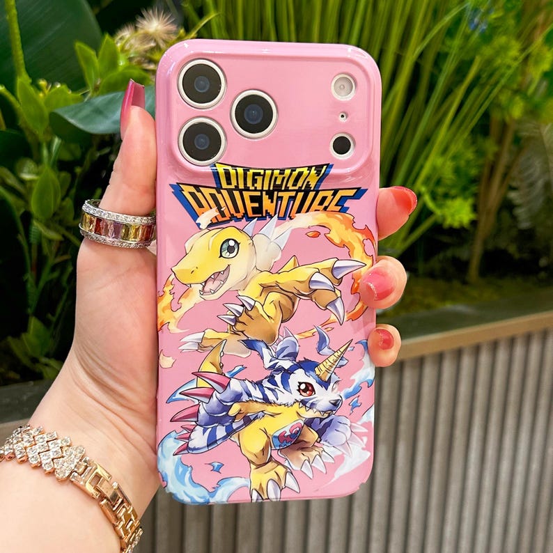May include: Pink phone case featuring the text "DIGIMON ADVENTURE" and illustrations of Digimon characters. The case has a glossy finish and cutouts for the camera and buttons.