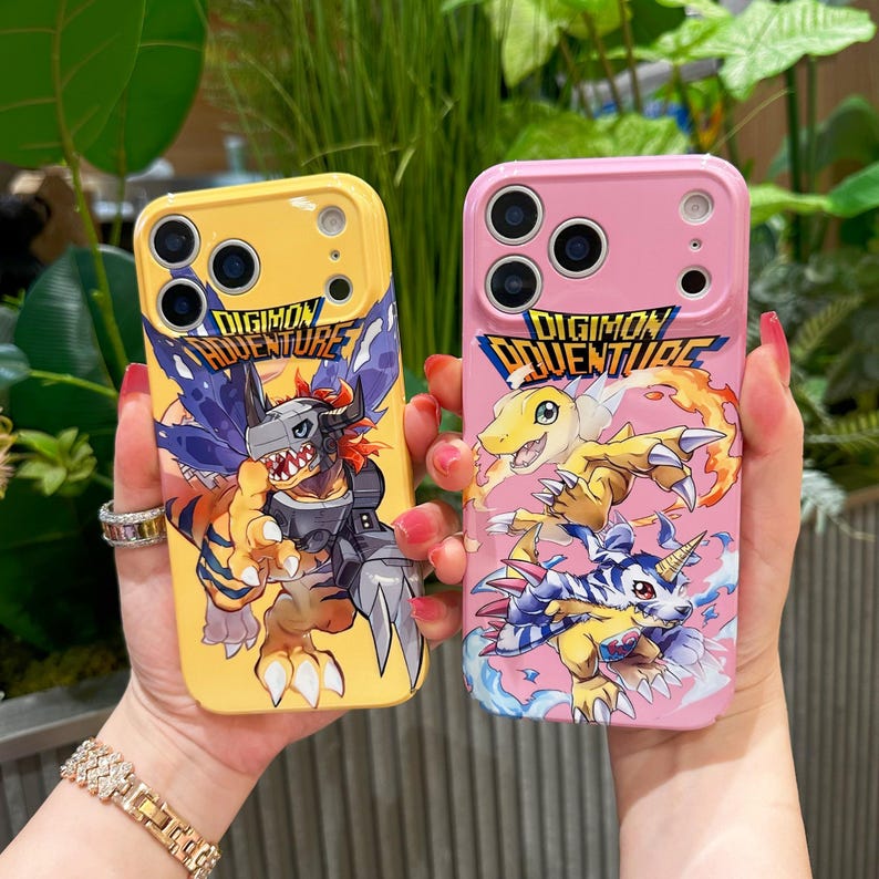 May include: Two phone cases, one yellow and one pink, featuring Digimon Adventure artwork. The yellow case displays a Digimon character with a gray and orange color scheme. The pink case shows three Digimon characters in yellow, orange, and blue.