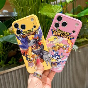 May include: Two phone cases, one yellow and one pink, featuring Digimon Adventure artwork. The yellow case displays a fierce orange and black Digimon with the text "DIGIMON ADVENTURE". The pink case shows a yellow Digimon and the text "DIGIMON ADVENTURE".