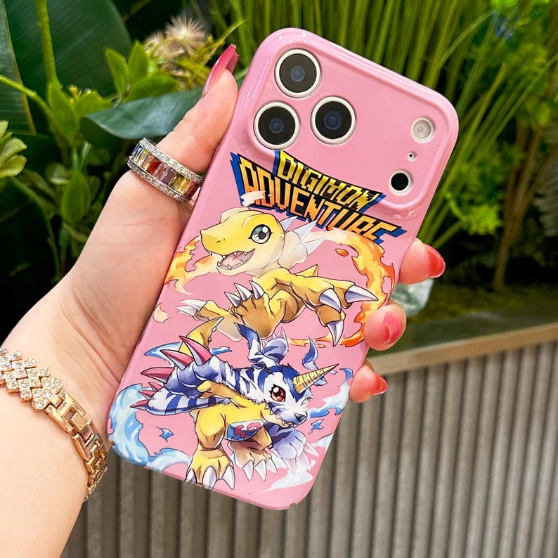 May include: Pink phone case featuring the text "Digimon Adventure" and cartoon characters. The case has a glossy finish and a design with yellow, blue, and white characters. The case has three camera cutouts.