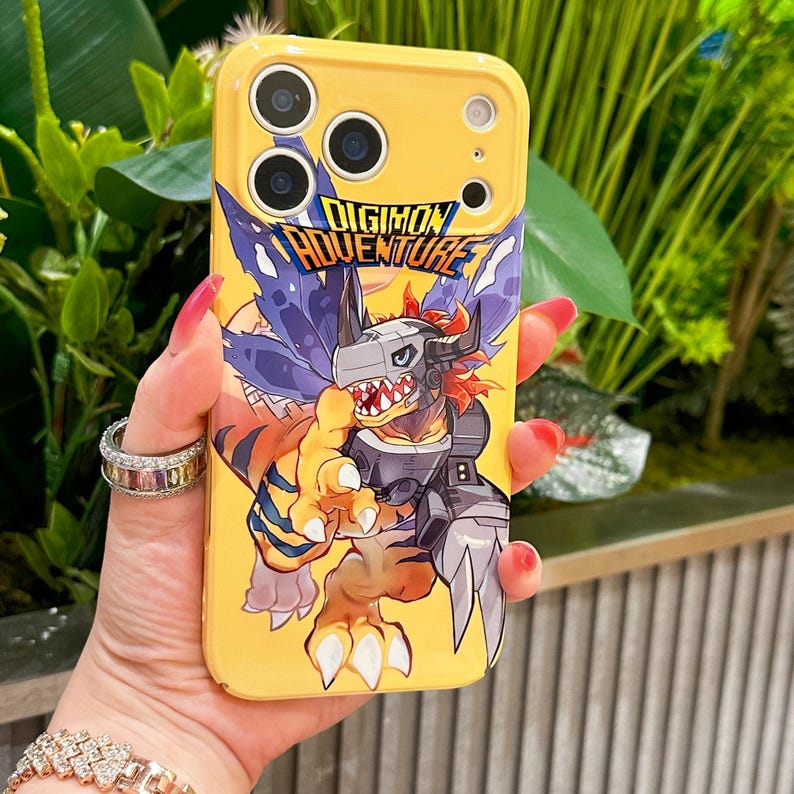 May include: Yellow phone case featuring a Digimon Adventure graphic. The case has a cartoon illustration of a monster with orange, gray, and blue colors. The text "DIGIMON ADVENTURE" is displayed in a blue and white font.