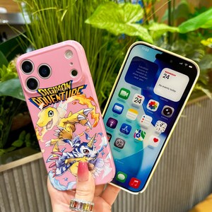 May include: Pink phone case featuring a Digimon Adventure graphic with yellow and blue characters. The case is held next to a phone displaying the date and time. The Digimon Adventure logo is visible on the case.