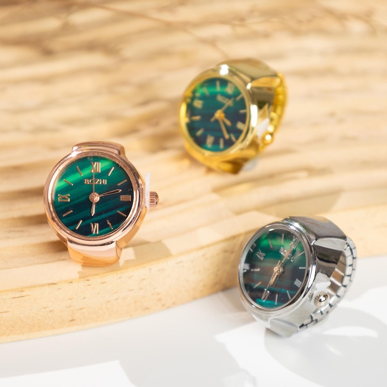 May include: Three ring watches with green faces and Roman numeral hour markers. One is gold-toned, one is rose gold-toned, and one is silver-toned. The watch faces have the word "BOZHI" printed on them.