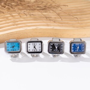May include: Four square-faced ring watches with silver-tone metal bands. The watch faces are white, black, and blue, with black numerals and hands. The watches display the time and have the word "Quartz" on the face.