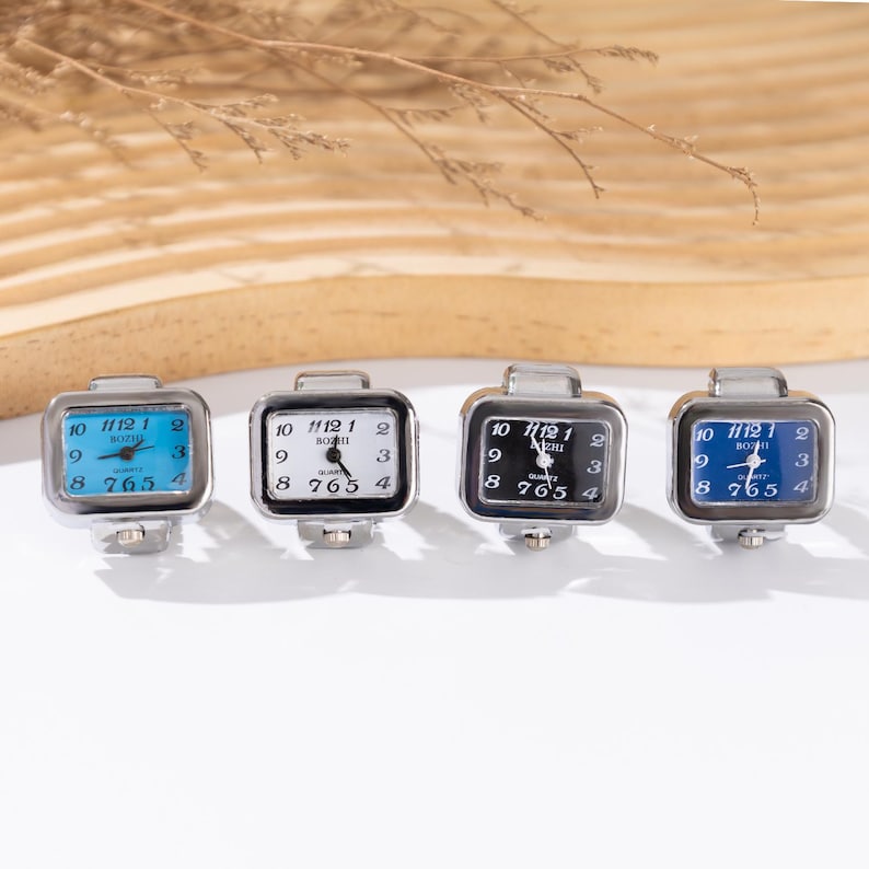 May include: Four square-faced ring watches with silver-tone metal bands. The watch faces are white, black, and blue, with black numerals and hands. The watches display the time and have the word "Quartz" on the face.