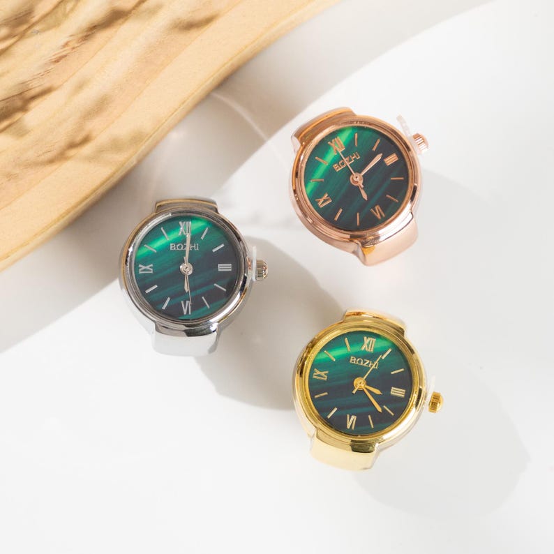 May include: Three ring watches with green faces and Roman numeral hour markers. The rings are silver, gold, and rose gold. The watch faces have the brand name "BOZHI" printed on them.