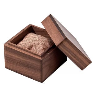 Puede incluir: Caja de reloj de madera marr&oacute;n oscuro con tapa con bisagras, parcialmente abierta. En el interior, un coj&iacute;n de color beige. La caja est&aacute; dise&ntilde;ada para guardar un reloj.