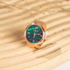 May include: A rose gold ring watch with a green face and Roman numeral hour markers. The watch face has the text "Bizhi". The ring watch is set on a light brown wooden surface.