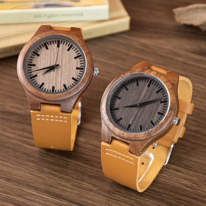 Puede incluir: Dos relojes de madera con correas de cuero marr&oacute;n claro. Las esferas de los relojes son de color marr&oacute;n claro y oscuro con manecillas y marcadores negros. Las cajas de los relojes son de madera. Los relojes se exhiben sobre una superficie de madera.
