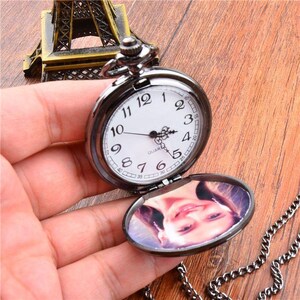 Puede incluir: Un reloj de bolsillo negro con una esfera blanca y n&uacute;meros negros. El reloj est&aacute; abierto, revelando una foto en el interior. Una cadena plateada est&aacute; adjunta. El reloj se sostiene en una mano.