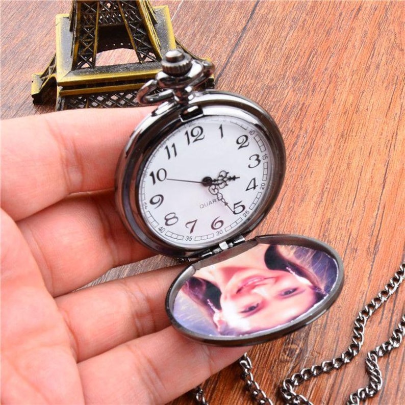 Puede incluir: Un reloj de bolsillo negro con una esfera blanca y n&uacute;meros negros. El reloj est&aacute; abierto, revelando una foto en el interior. Una cadena plateada est&aacute; adjunta. El reloj se sostiene en una mano.