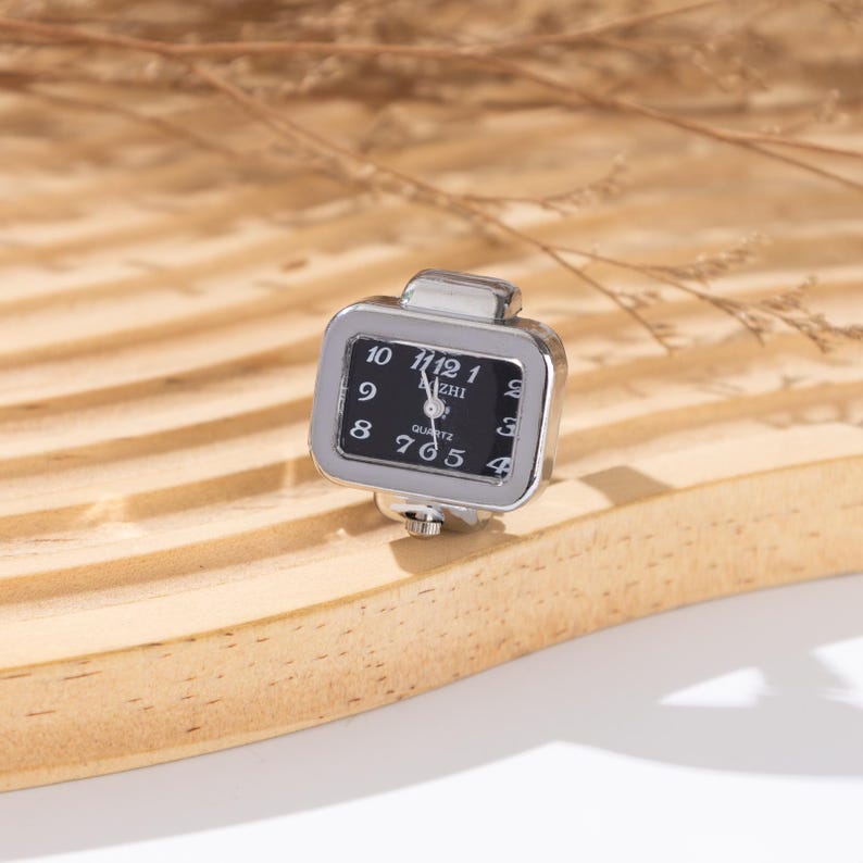 May include: A silver-toned ring watch with a square face. The watch face is black with white numerals and hands, displaying the time. The watch has the word "Quartz" printed on the face. The ring is set on a wooden surface.