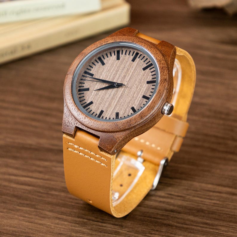Puede incluir: Reloj de pulsera de madera con correa de cuero marr&oacute;n claro. La esfera del reloj es de madera con marcas y agujas negras. La caja del reloj es de color marr&oacute;n oscuro. El reloj se muestra sobre una superficie de madera.