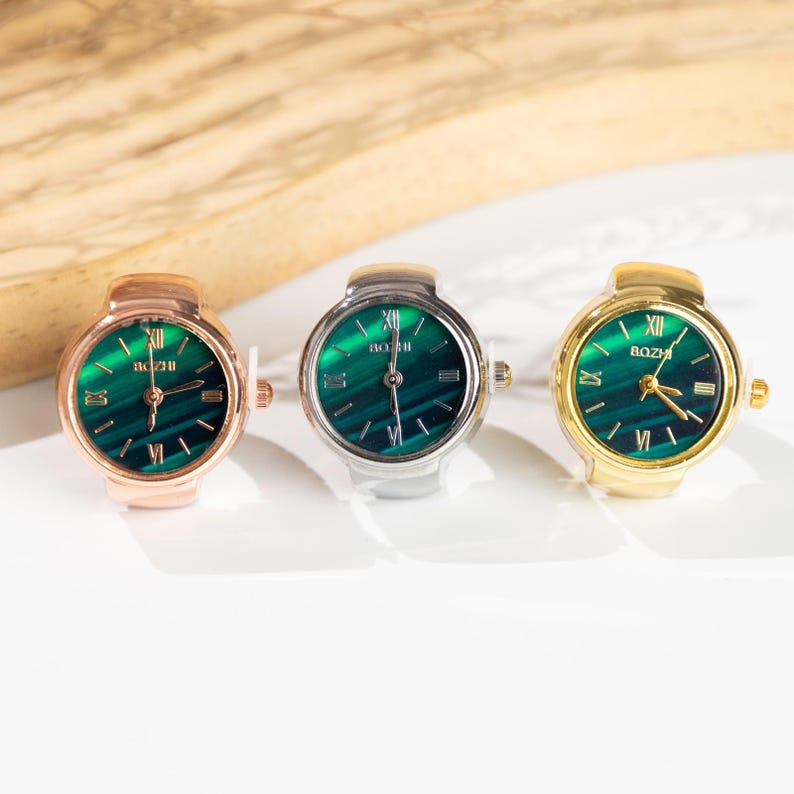 May include: Three ring watches with green watch faces and gold, silver, and rose gold bands. The watch faces have Roman numerals and the brand name "BOZI". The watches are displayed on a white surface.