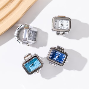 May include: Four square-faced ring watches with silver-tone metal bands. The watch faces are white or blue with black numerals and hands. The watch rings are a unique accessory.