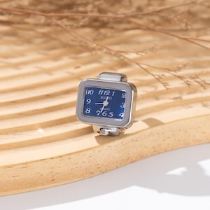 May include: A silver-toned ring watch with a square blue face. The watch has white numerals and hands, and the brand name "BOZHI" is printed on the face. The ring is set on a light brown wooden surface.
