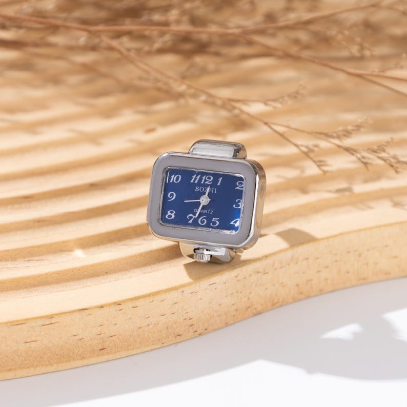May include: A silver-toned ring watch with a square blue face. The watch has white numerals and hands, and the brand name "BOZHI" is printed on the face. The ring is set on a light brown wooden surface.