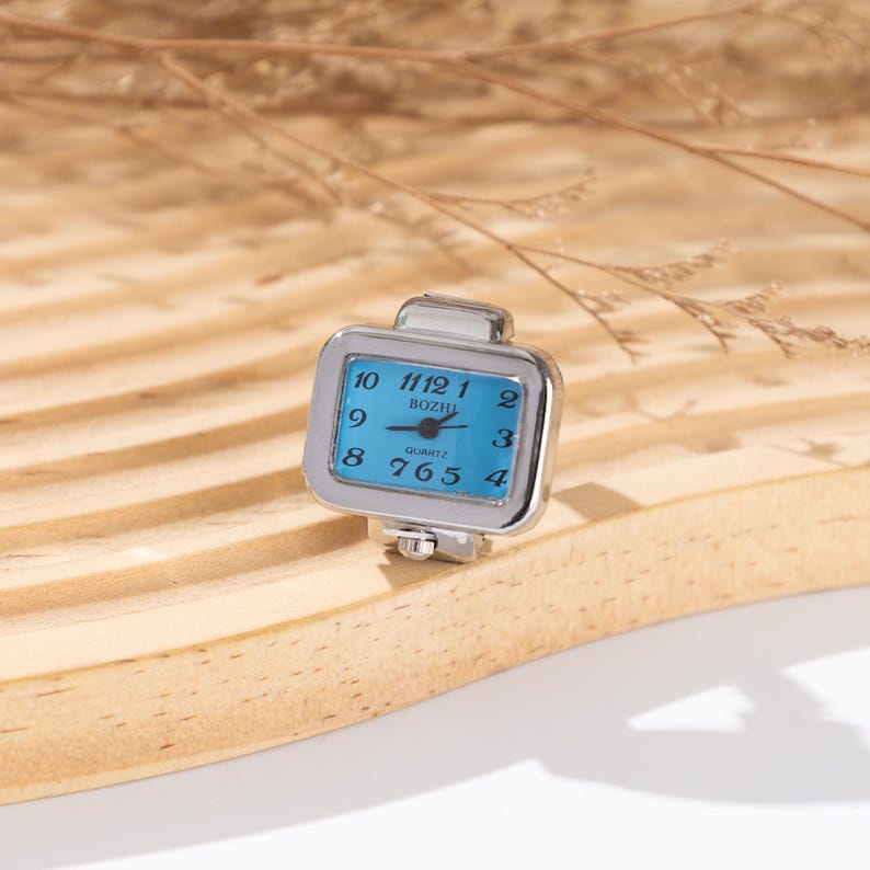 May include: A silver-toned ring watch with a square, light blue face. The watch face displays the numbers 1 through 12, with the brand name "BOZHI" printed in the center. The watch is a quartz movement.