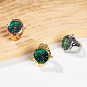 May include: Three ring watches with green watch faces and gold, rose gold, and silver bands. The watch faces have Roman numerals and gold hands. The rings are displayed on a light-colored wooden surface.
