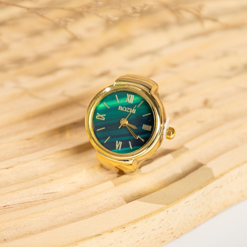 May include: A gold-toned ring watch with a green and blue face. The watch face has Roman numerals and the brand name "BOZHI". The ring is set on a light brown wooden surface.