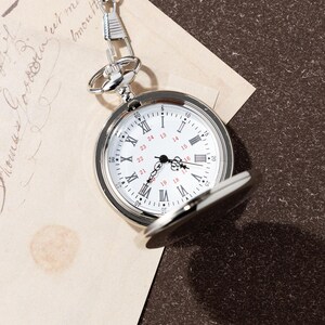 Puede incluir: Un reloj de bolsillo plateado con esfera blanca, manecillas negras y n&uacute;meros romanos. El reloj est&aacute; parcialmente abierto, revelando la hora. Tiene una cadena y est&aacute; sobre un fondo de papel vintage.