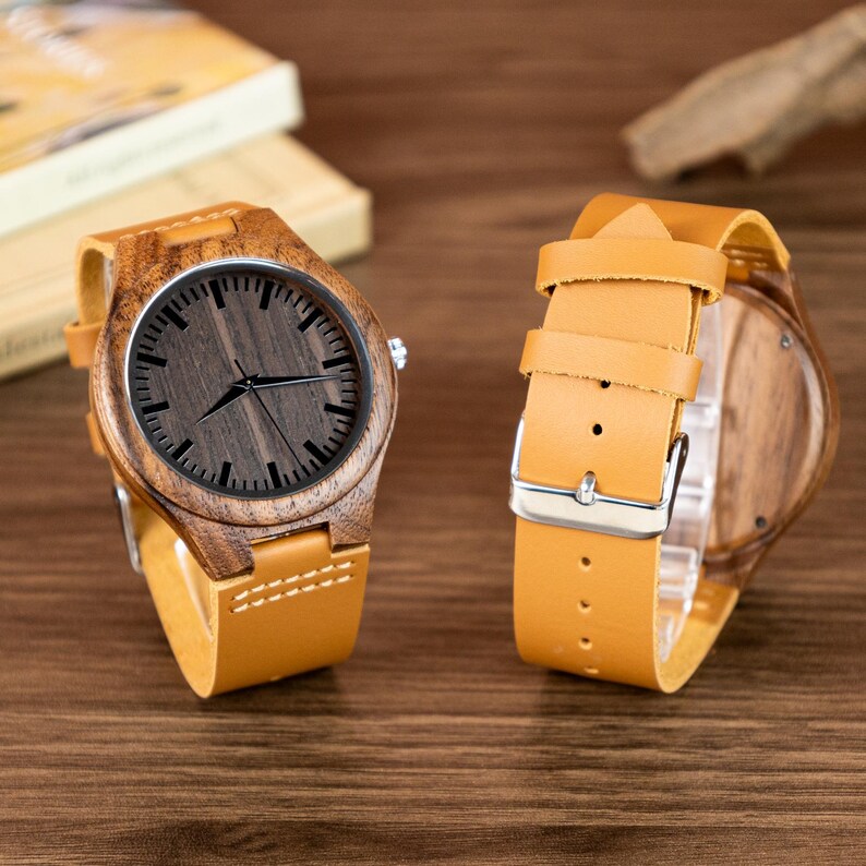 Puede incluir: Dos relojes de pulsera de madera con esferas de madera marr&oacute;n y correas de cuero color canela. Las esferas tienen marcas y manecillas negras. Las cajas de los relojes son de madera. Las correas de cuero tienen hebillas plateadas.