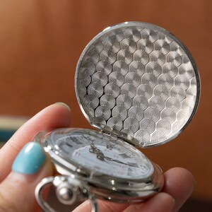 Puede incluir: Un reloj de bolsillo plateado con la tapa abierta, que revela una esfera blanca con n&uacute;meros romanos negros y manecillas. La tapa del reloj tiene un dise&ntilde;o texturizado de c&iacute;rculos conc&eacute;ntricos. El reloj se sostiene en una mano.