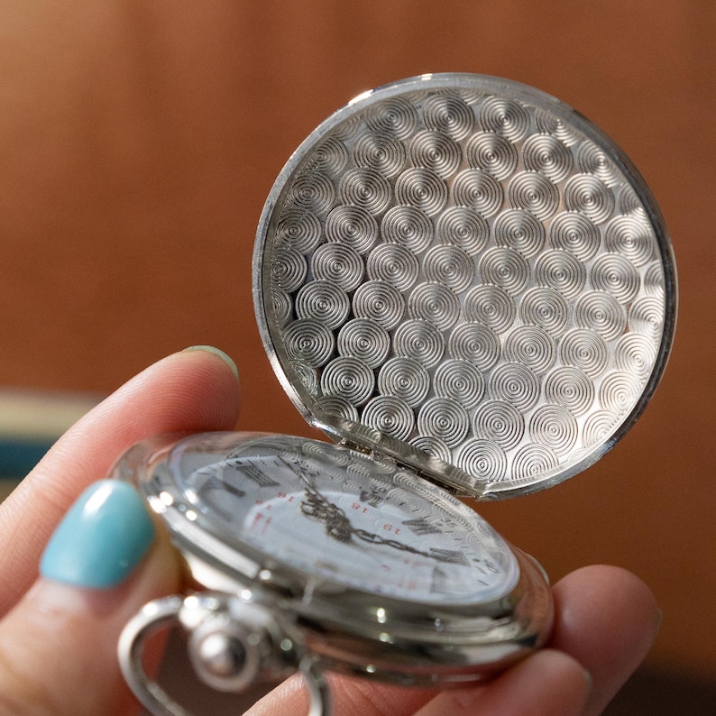 Puede incluir: Un reloj de bolsillo plateado con la tapa abierta, que revela una esfera blanca con n&uacute;meros romanos negros y manecillas. La tapa del reloj tiene un dise&ntilde;o texturizado de c&iacute;rculos conc&eacute;ntricos. El reloj se sostiene en una mano.