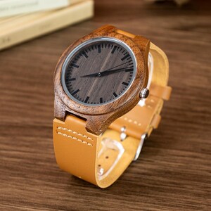 Puede incluir: Un reloj de pulsera de madera con una esfera de madera oscura y una correa de cuero marr&oacute;n claro. El reloj tiene un bisel plateado y agujas negras. El reloj se muestra sobre una superficie de madera.